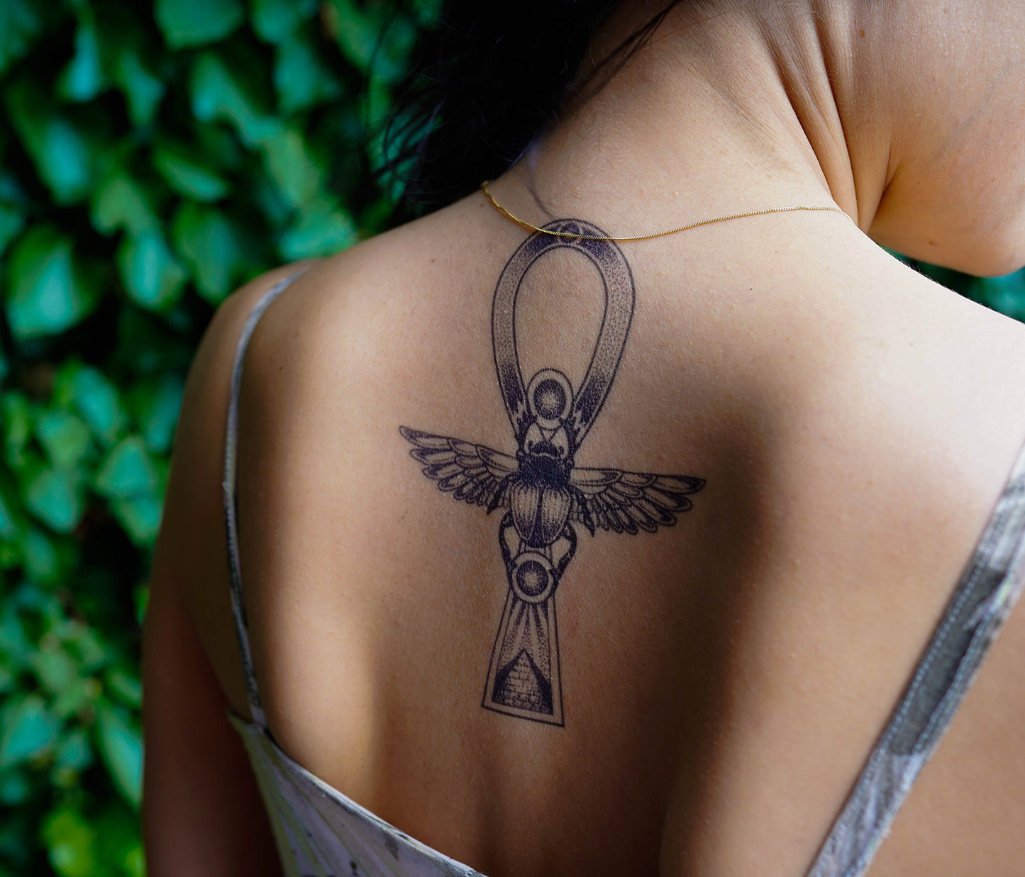 A person with a detailed temporary tattoo of an Ankh symbol with wings and a beetle on their upper back, reflecting Ancient Egypt